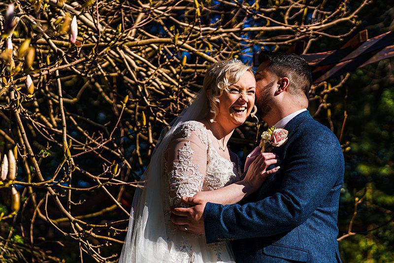A bride and groom share a joyful moment under a tree with bare branches. The bride, in a lace wedding dress, laughs heartily while the groom, in a dark suit, smiles and leans in to kiss her forehead. The scene is warmly lit by sunlight. Image by Documentary Wedding Photographer Lewis Bishop Photography.
