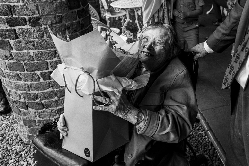 An elderly person in a wheelchair smiles while holding a large bouquet of flowers wrapped in paper. Image by Documentary Wedding Photographer Lewis Bishop Photography.