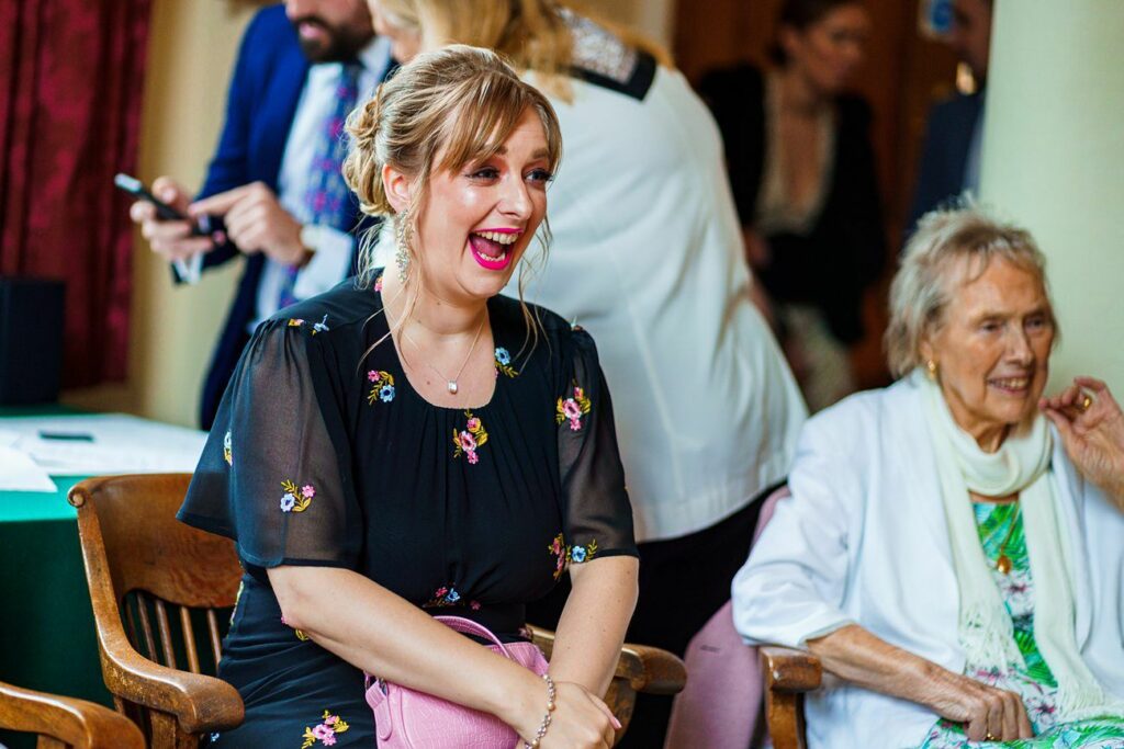 A woman in a black dress with floral patterns, laughing and sitting on a wooden chair. An elderly woman in a white cardigan sits to her left. A few other people are visible in the background, engaging in conversation. The atmosphere seems joyful and lively. Image by Documentary Wedding Photographer Lewis Bishop Photography.