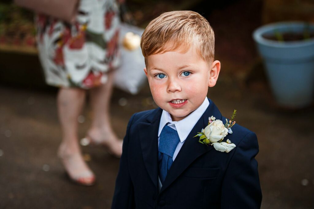 A young boy with blond hair and blue eyes, dressed in a navy suit with a blue tie and a white flower boutonniere, smiles at the camera. In the blurred background, an adult in a floral dress is partially visible, standing outdoors with flower petals on the ground. Image by Documentary Wedding Photographer Lewis Bishop Photography.