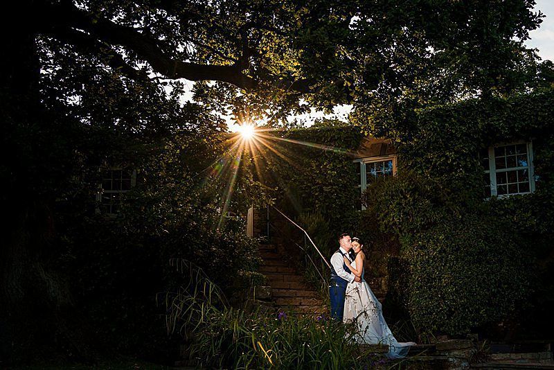 A bride and groom embrace on a stone staircase covered in vines and lush greenery. The sun is setting, casting a warm glow and beams of light through the trees above them, creating a romantic atmosphere. The bride's veil flows gracefully down the steps. Image by Documentary Wedding Photographer Lewis Bishop Photography.