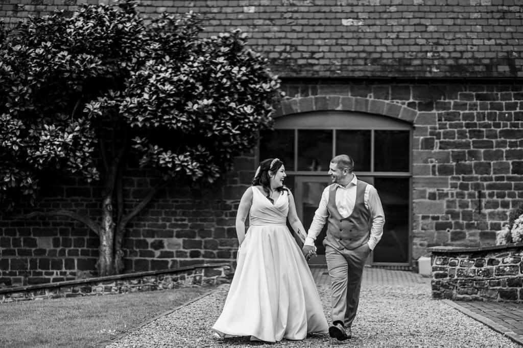 A bride and groom walk hand in hand on a gravel path beside a brick building with a tree in the background. Image by Documentary Wedding Photographer Lewis Bishop Photography.