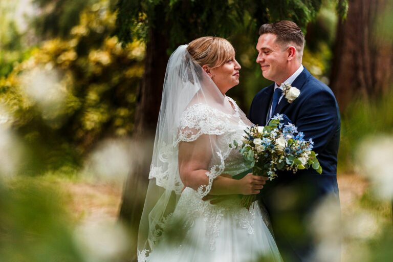 A bride and groom gaze lovingly at each other on their wedding day. The bride is holding a bouquet of flowers, dressed in a white lace gown and veil, while the groom is in a dark suit with a boutonnière. They are surrounded by greenery and trees. Image by Documentary Wedding Photographer Lewis Bishop Photography.