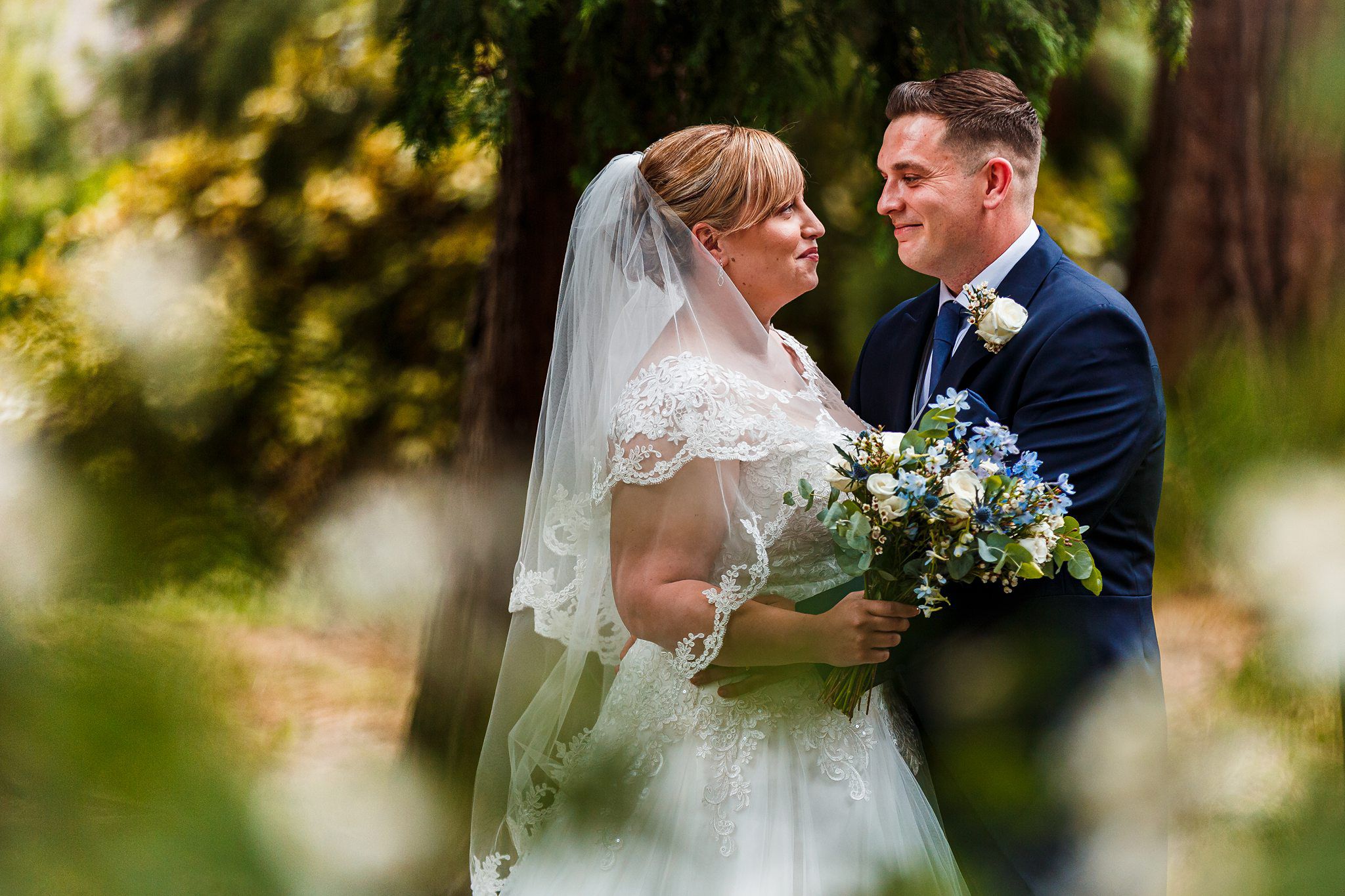 A bride and groom gaze lovingly at each other on their wedding day. The bride is holding a bouquet of flowers, dressed in a white lace gown and veil, while the groom is in a dark suit with a boutonnière. They are surrounded by greenery and trees. Image by Documentary Wedding Photographer Lewis Bishop Photography.