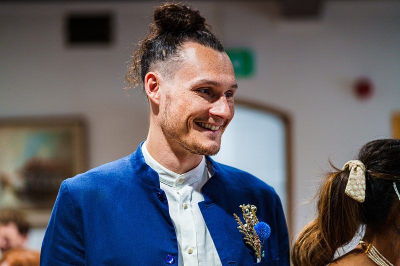 A man with a top knot hairstyle, wearing a blue jacket over a white shirt, and a flower pinned to his jacket, smiles while looking slightly to his right. Image by Documentary Wedding Photographer Lewis Bishop Photography.