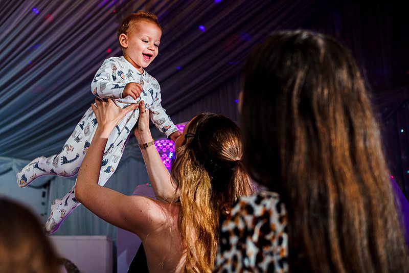 A smiling baby in a onesie with animal prints is being lifted in the air by an adult with long brown hair, while another person with long hair watches from behind. The background shows a room with draped fabric and colorful lights. Image by Documentary Wedding Photographer Lewis Bishop Photography.