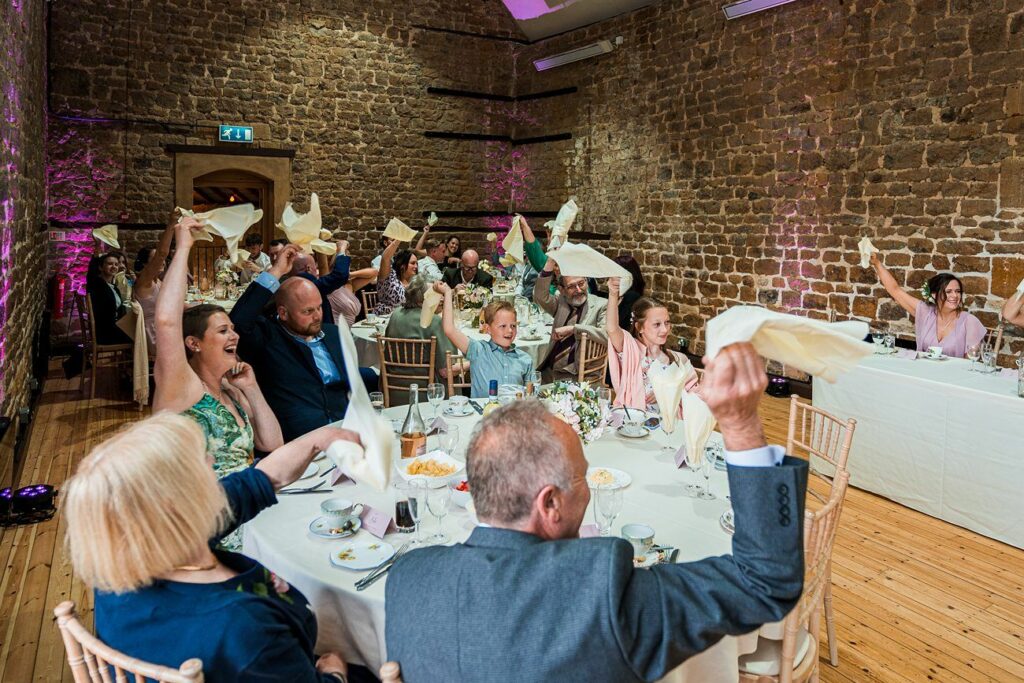 Guests seated at round tables wave napkins in the air during a lively moment at an indoor event, with a stone wall backdrop and ambient lighting. Image by Documentary Wedding Photographer Lewis Bishop Photography.
