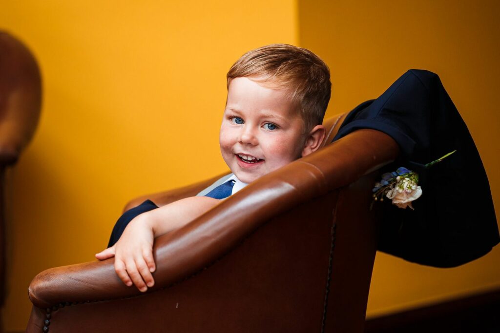 A young boy with light brown hair, wearing a dark blue suit, is sitting in a brown leather chair and smiling. He has a white shirt on and a boutonniere with flowers pinned to his suit. The background is a warmly lit, golden yellow wall. Image by Documentary Wedding Photographer Lewis Bishop Photography.