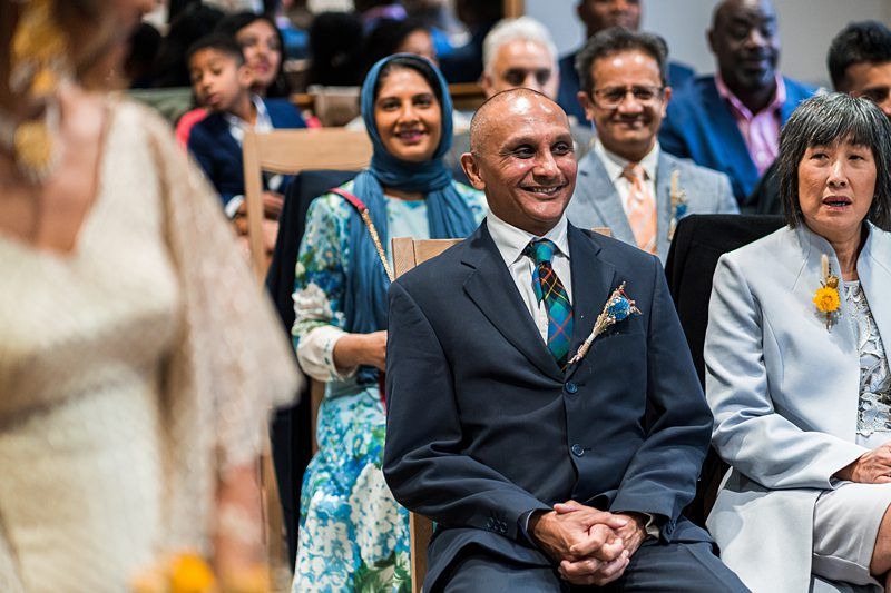 People are seated in a ceremony, with a smiling man in a suit and tie in the forefront and other attendees, some dressed formally, visible in the background. Image by Documentary Wedding Photographer Lewis Bishop Photography.