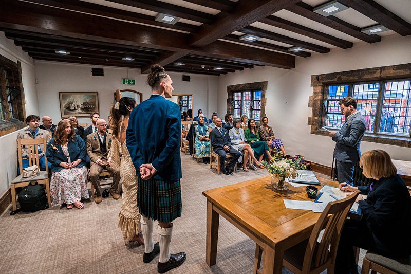 A couple stands at the front of a room facing an officiant during a wedding ceremony, with guests seated behind them and two people at a table to the side. Image by Documentary Wedding Photographer Lewis Bishop Photography.