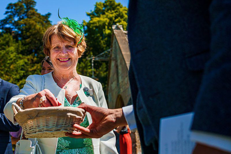 An older woman wearing a green dress and green fascinator smiles while placing something into a wicker basket held by a person out of focus in the foreground. They are outdoors with trees and sunlight in the background. Image by Documentary Wedding Photographer Lewis Bishop Photography.