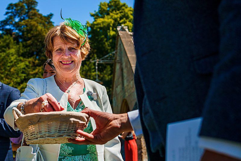 An elderly woman wearing a green outfit and green fascinator is smiling while placing something in a basket held by another person. She is outdoors, with trees and a building in the background on a sunny day. Image by Documentary Wedding Photographer Lewis Bishop Photography.