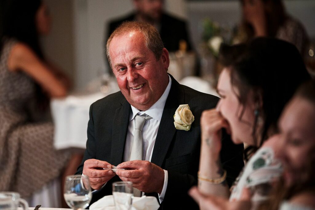 A man in a suit with a white rose boutonniere sits at a table, smiling warmly at someone off-camera. He is holding a small object in his hands. The atmosphere appears to be that of a formal social gathering, with other attendees in the background. Image by Documentary Wedding Photographer Lewis Bishop Photography.