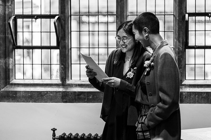 Two people stand in front of a window. One is reading a document, and both are smiling. The background shows a wall with three rectangular, stained glass windows. Image by Documentary Wedding Photographer Lewis Bishop Photography.