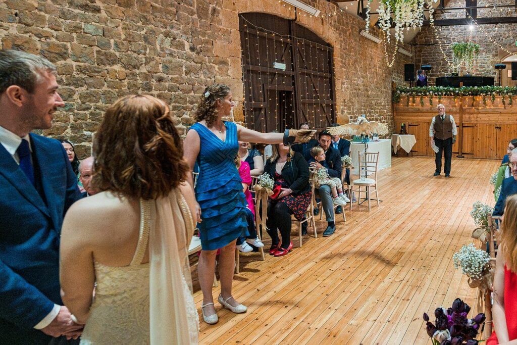 A bride and groom stand at the front of a wedding ceremony in a rustic hall. A woman in a blue dress next to the groom holds a falcon, drawing attention from seated guests. Image by Documentary Wedding Photographer Lewis Bishop Photography.