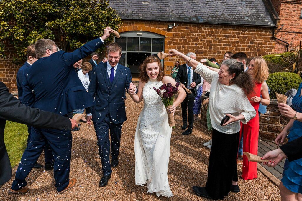A newlywed couple walks down a path lined with guests who are throwing confetti. The bride holds a bouquet of dark red flowers. The groom wears a blue suit, and both are smiling. Image by Documentary Wedding Photographer Lewis Bishop Photography.