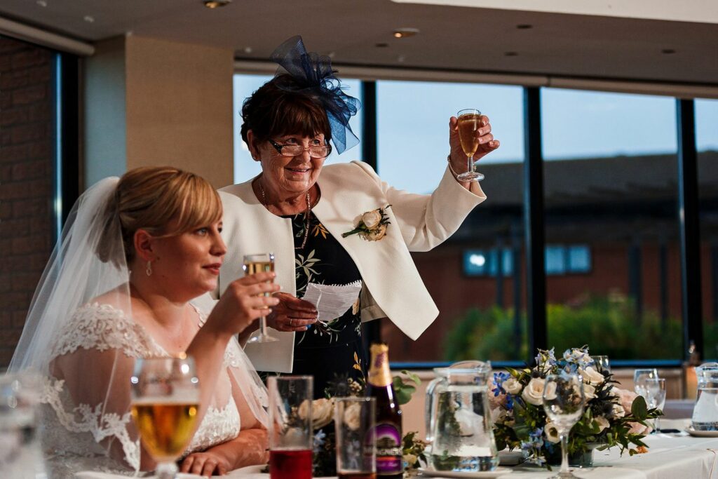 An older woman in a white jacket with a floral brooch is giving a toast while holding a glass of wine. A bride in a white wedding dress and veil sits beside her, also holding up a glass. They are at a wedding reception with a decorated table in front of them. Image by Documentary Wedding Photographer Lewis Bishop Photography.