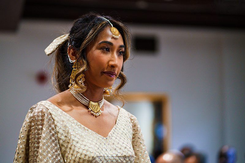 A woman wearing traditional South Asian attire, including jewelry and a headpiece, looks ahead. She has wavy hair and a ribbon tied at the back. She is indoors in a softly lit environment. Image by Documentary Wedding Photographer Lewis Bishop Photography.