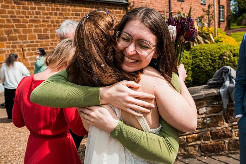 Two women are hugging outdoors while smiling. One is holding a bouquet of dark flowers. Other people and brick buildings are visible in the background. Image by Documentary Wedding Photographer Lewis Bishop Photography.