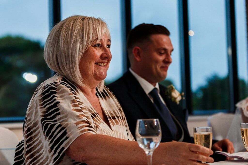 A smiling woman with short, light-colored hair sits at a table with a glass of champagne in front of her. She wears a patterned blouse. Next to her, a man in a suit and tie, also smiling, has a glass of champagne. They are indoors with a window in the background. Image by Documentary Wedding Photographer Lewis Bishop Photography.