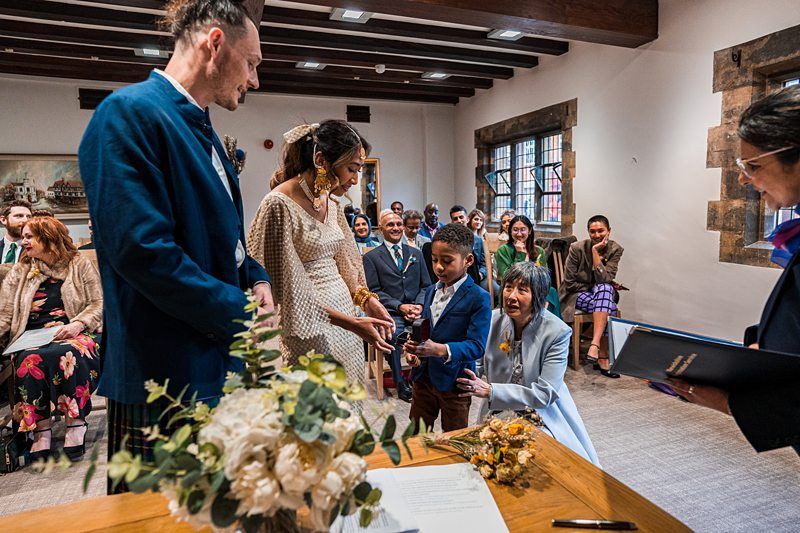 A young boy exchanges rings with a couple, dressed in wedding attire, in a small room with guests looking on and a bouquet on the table in the foreground. Image by Documentary Wedding Photographer Lewis Bishop Photography.