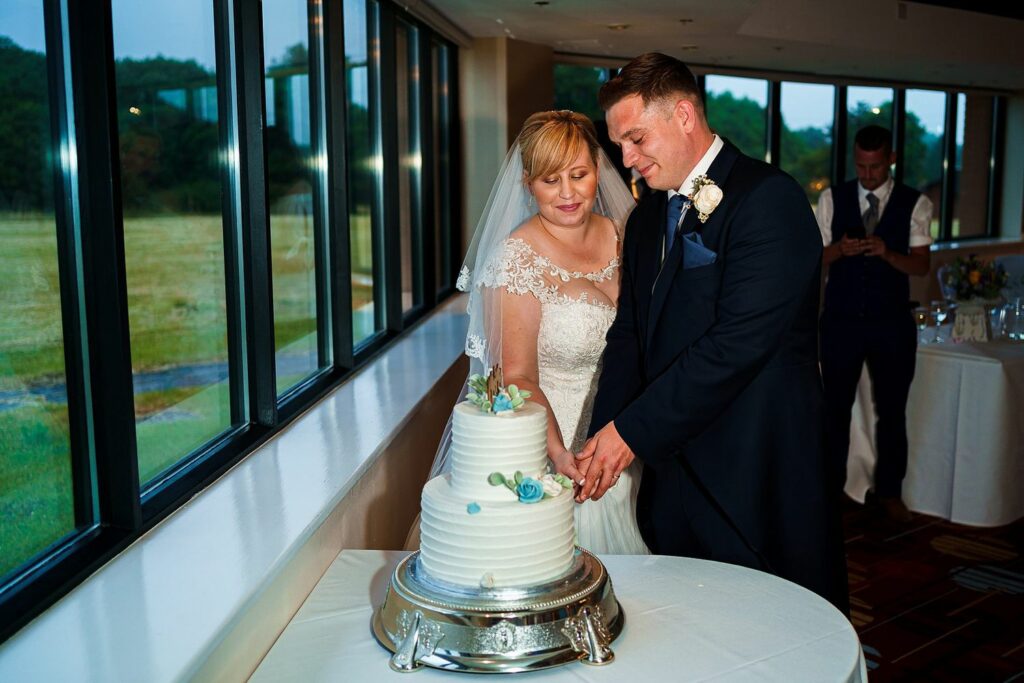 A bride and groom, in a white wedding dress and dark suit respectively, are cutting a white tiered wedding cake adorned with blue flowers. They are inside a room with large windows overlooking a green landscape. Another person in formal attire is in the background. Image by Documentary Wedding Photographer Lewis Bishop Photography.