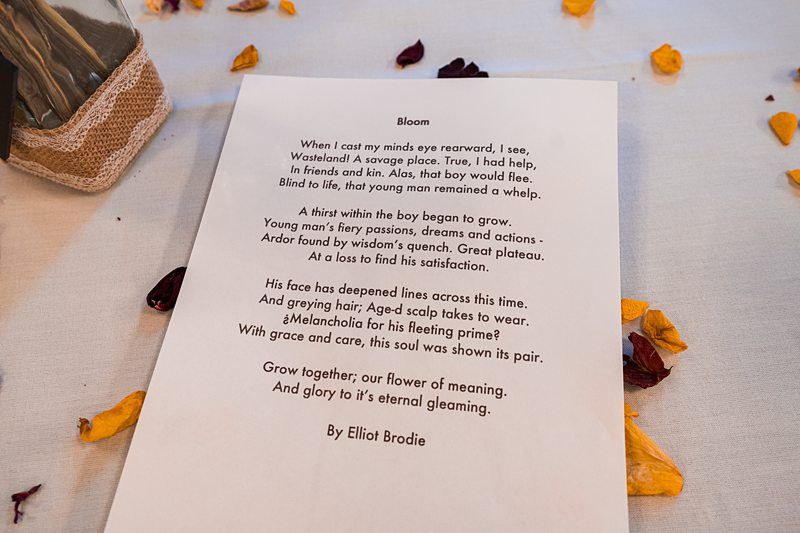 A printed poem titled “Bloom” by Elliot Brodie lies on a table, surrounded by scattered yellow and red flower petals. Image by Documentary Wedding Photographer Lewis Bishop Photography.