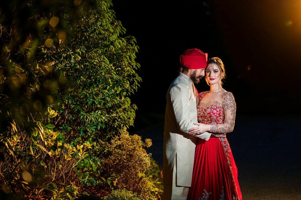 A couple stands close together in traditional attire; the man wears a cream-colored outfit and a red turban, while the woman wears a red dress with intricate gold patterns. They are illuminated against a dark background with foliage visible on the left side. Image by Documentary Wedding Photographer Lewis Bishop Photography.