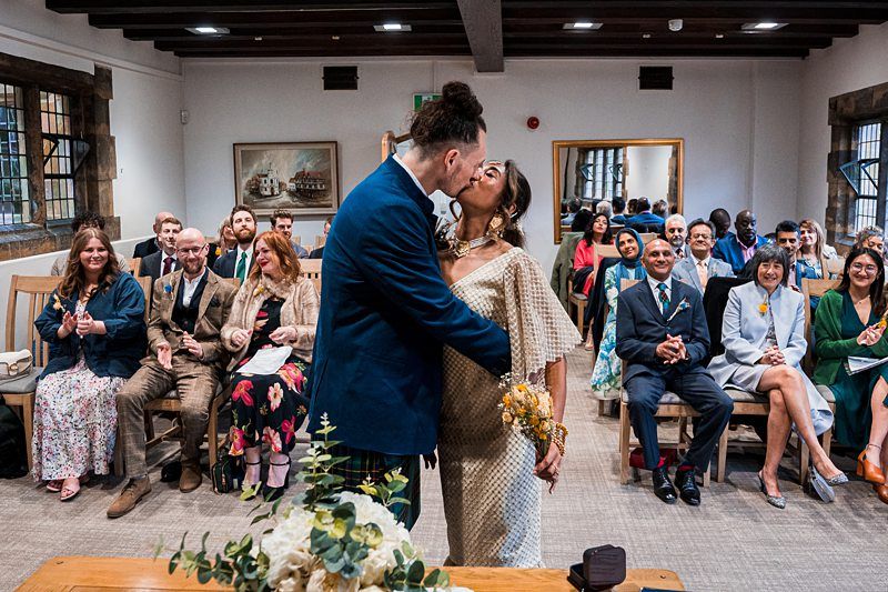 A bride and groom share a kiss in front of seated guests during their wedding ceremony in a small room. The guests are smiling and clapping. A bouquet and floral arrangement are visible in the foreground. Image by Documentary Wedding Photographer Lewis Bishop Photography.
