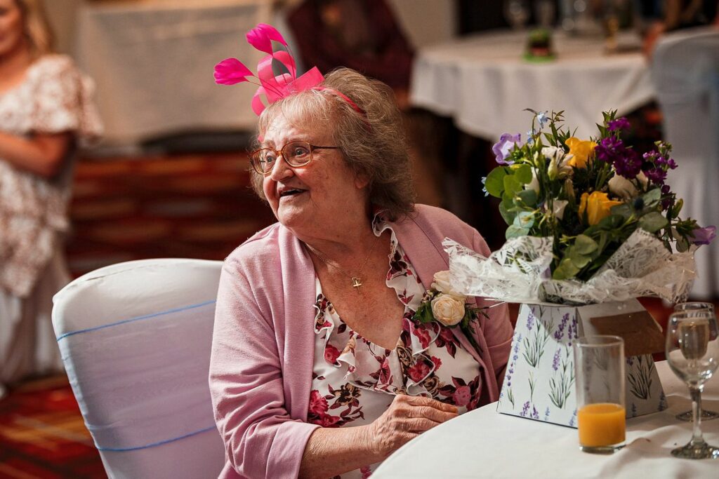 An elderly woman with a pink headpiece sits at a table, smiling. She wears glasses and a floral dress with a pink cardigan. In front of her is a gift wrapped in floral paper and a vase filled with flowers. There are people and tables in the background. Image by Documentary Wedding Photographer Lewis Bishop Photography.