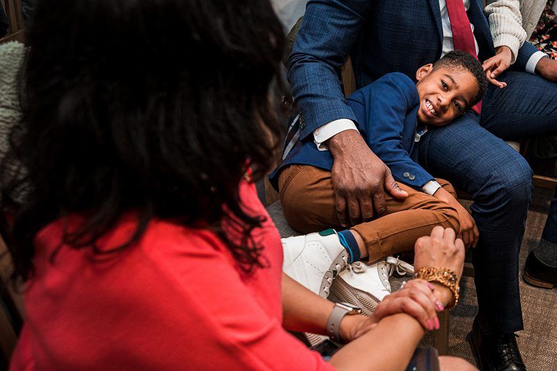 A young boy, dressed in a blue blazer and brown pants, smiles while sitting on a man's lap. A woman in a red shirt and white sneakers is seated nearby, looking at the boy. Image by Documentary Wedding Photographer Lewis Bishop Photography.