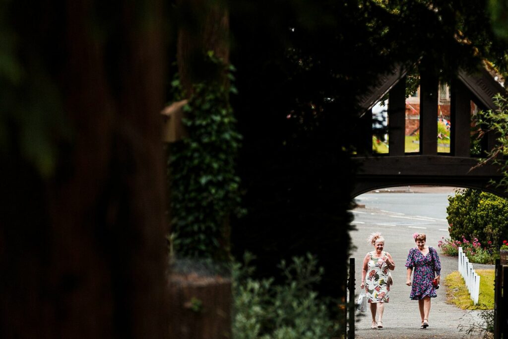 Two women in floral dresses walk along a path bordered by trees towards a wooden archway. The path is lined with greenery and leads to a street in the background. Both women appear to be engaged in conversation. Image by Documentary Wedding Photographer Lewis Bishop Photography.