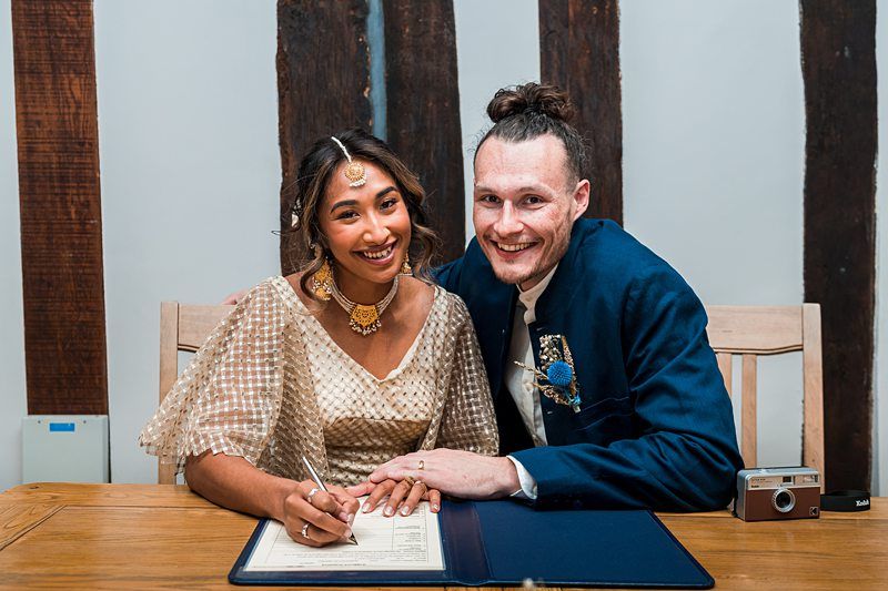 A couple dressed in formal attire smiles at the camera while the woman signs a document on a wooden table. Image by Documentary Wedding Photographer Lewis Bishop Photography.