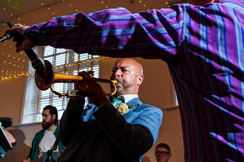 A man in a blue suit plays the trumpet passionately at an indoor event. Another man in a purple striped shirt adjusts a microphone in front of him. There are string lights in the background and people watching attentively. Image by Documentary Wedding Photographer Lewis Bishop Photography.