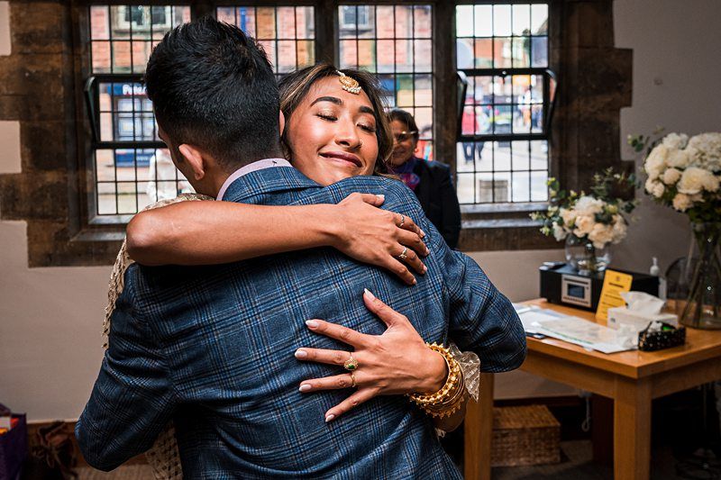 A woman wearing gold jewelry hugs a man in a checkered jacket inside a room with large windows and floral decorations. Image by Documentary Wedding Photographer Lewis Bishop Photography.