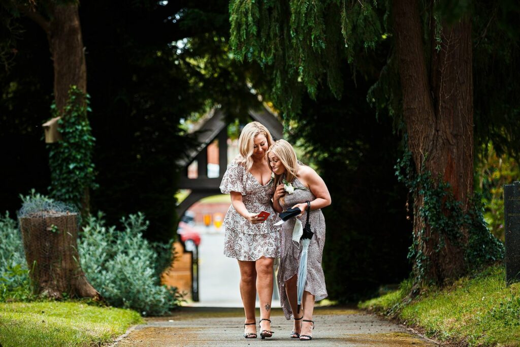 Two women in summer dresses walk together down a tree-lined path. One points at something on her phone while the other looks on, smiling. A blurred building and greenery are in the background, creating a serene, outdoor atmosphere. Image by Documentary Wedding Photographer Lewis Bishop Photography.
