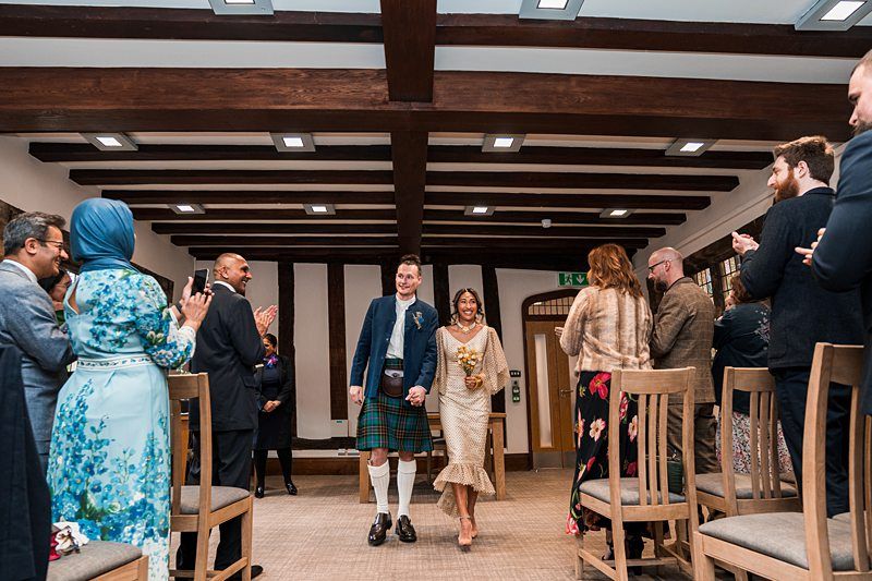 A couple walks down the aisle holding hands at their wedding ceremony, while guests on either side clap and smile. The groom is in a kilt and jacket, and the bride is in a light-colored dress holding flowers. Image by Documentary Wedding Photographer Lewis Bishop Photography.