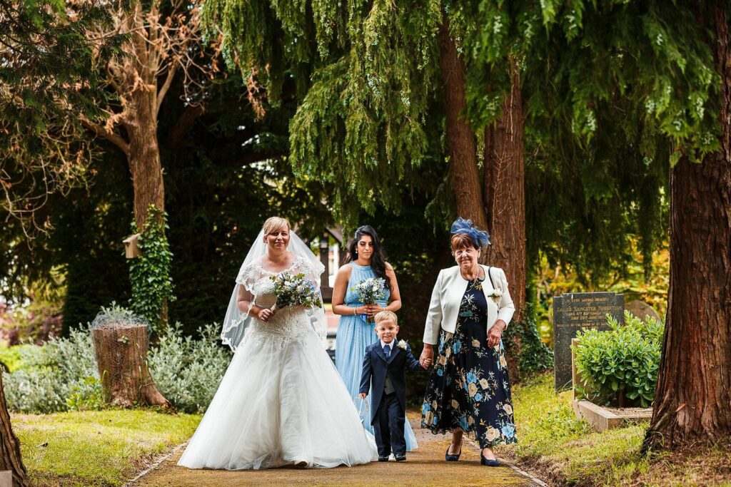 A bride in a white wedding dress walks down a lush garden path, holding a bouquet and arm-in-arm with an older woman in a floral dress and white jacket. They are accompanied by a young child in a suit and a bridesmaid in a light blue dress holding a bouquet. Image by Documentary Wedding Photographer Lewis Bishop Photography.