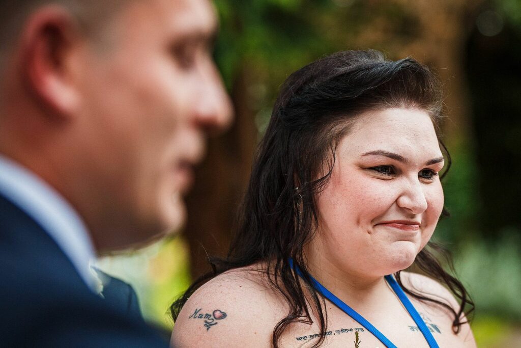 A woman with long dark hair and visible tattoos on her shoulders smiles while looking at a person standing next to her. The background is blurred, featuring some greenery. The person beside her is slightly out of focus. Image by Documentary Wedding Photographer Lewis Bishop Photography.