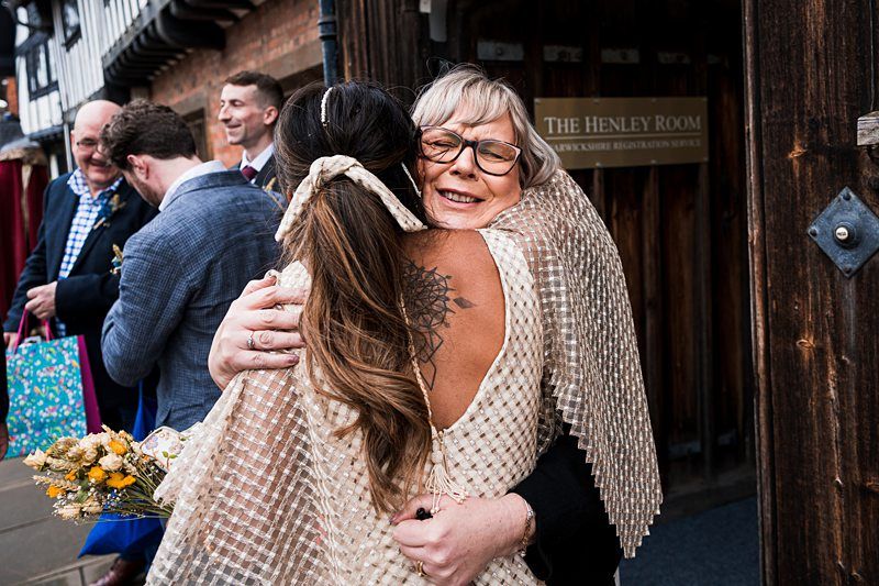 Two women embrace outside a building labeled "The Henley Room." One woman has a floral tattoo on her back and the other wears glasses and a beige shawl. Other people are chatting in the background. Image by Documentary Wedding Photographer Lewis Bishop Photography.