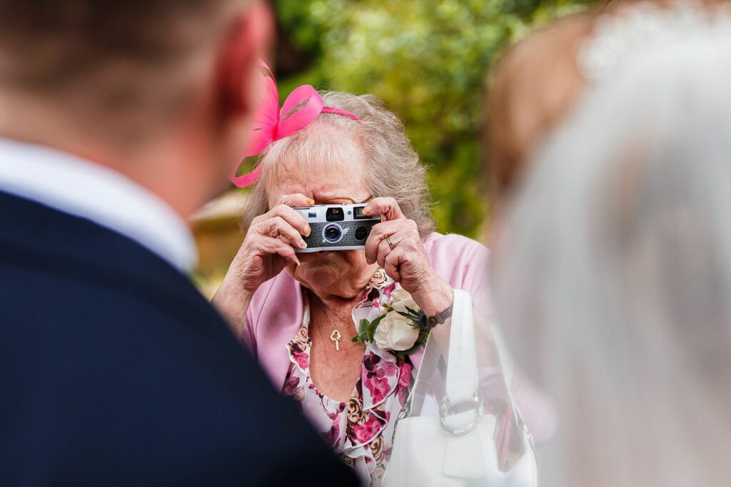 An elderly woman wearing a pink fascinator takes a photo with a compact camera at an outdoor event. She is partially obscured by two people, one in black attire and the other in white, suggesting a wedding scene. Lush greenery is visible in the background. Image by Documentary Wedding Photographer Lewis Bishop Photography.