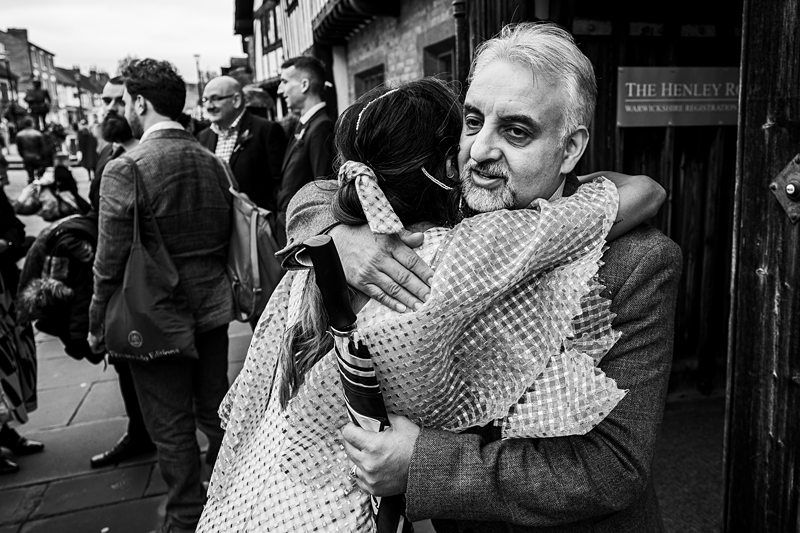 A woman in a patterned dress embraces an older man with greying hair and a beard on a busy street. Other people are visible in the background near a sign reading "The Henley Row. Image by Documentary Wedding Photographer Lewis Bishop Photography.