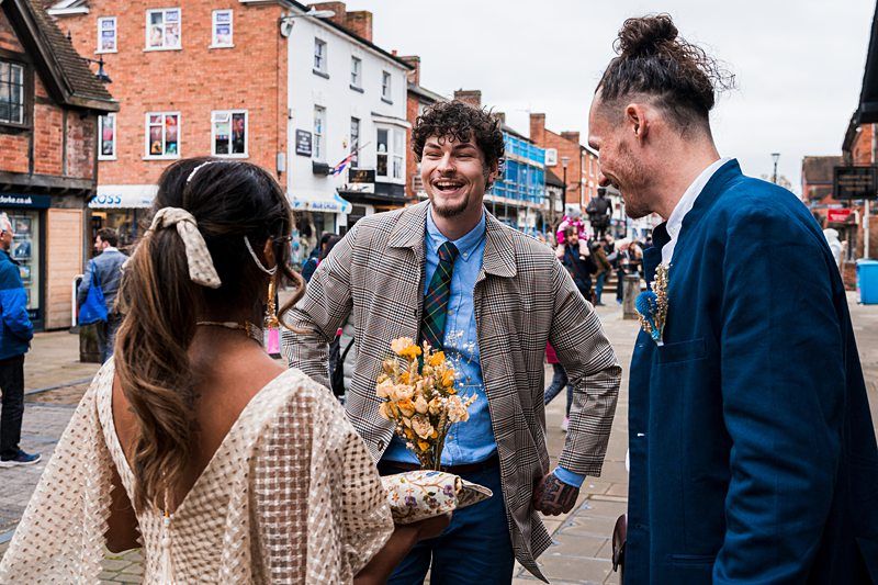 Three people stand and chat on a lively street lined with shops. One woman holds a small bouquet of flowers while two men, one wearing a tie and coat and the other in a blue jacket, smile and engage with her. Image by Documentary Wedding Photographer Lewis Bishop Photography.