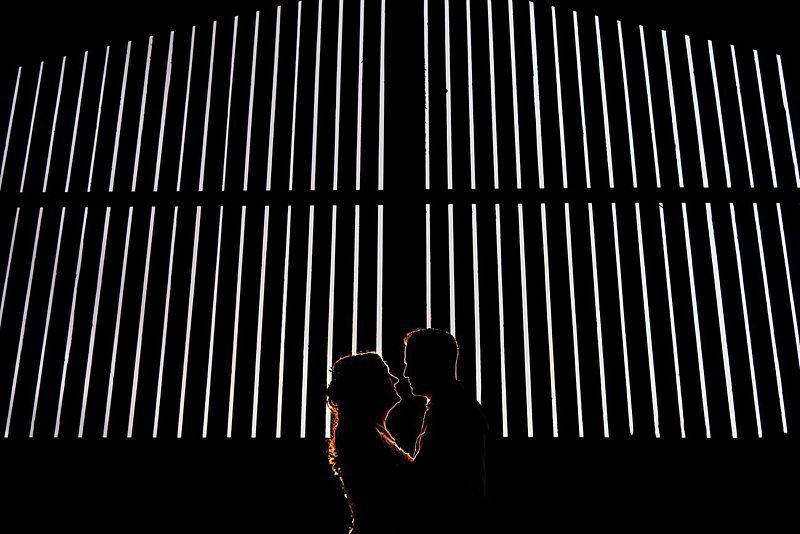 Silhouetted couple facing each other in an embrace against a backdrop of vertical white light bars in a dark setting. Image by Documentary Wedding Photographer Lewis Bishop Photography.