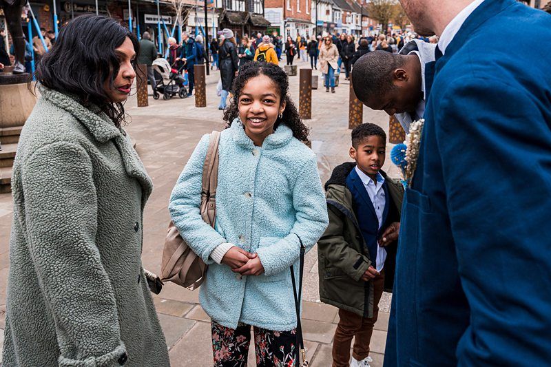 A family stands together in an outdoor plaza. The girl in a light blue coat is smiling, the boy in a dark coat looks on, and the two adults are engaged in conversation. People walk in the background. Image by Documentary Wedding Photographer Lewis Bishop Photography.