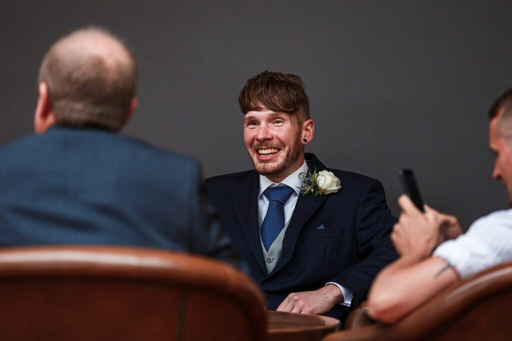 A man in a suit with a flower boutonniere is smiling and sitting at a table. He is facing two other men; one is bald and wearing a suit, and the other is holding a smartphone. The background is neutral and dark. Image by Documentary Wedding Photographer Lewis Bishop Photography.