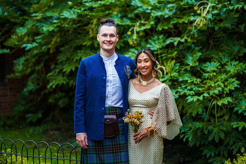 A couple stands together outdoors, with the man wearing a suit jacket and kilt and the woman wearing a traditional dress, both smiling in front of lush greenery. Image by Documentary Wedding Photographer Lewis Bishop Photography.