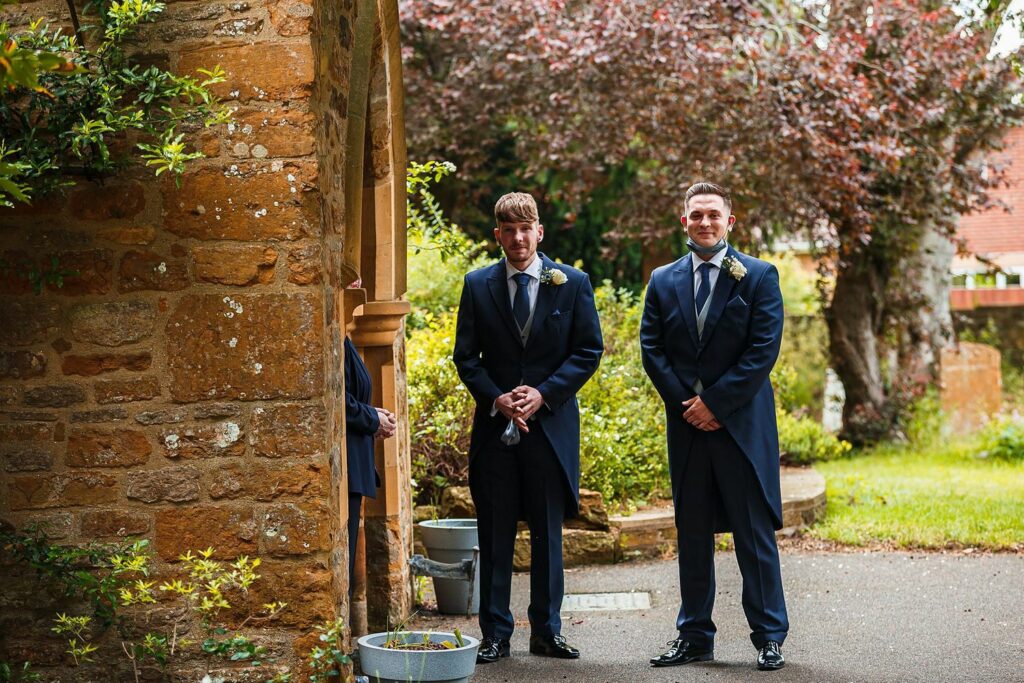 Two men dressed in dark blue suits with white boutonnieres stand next to a stone building. One man holds a paper and a water bottle. The scene is set outdoors with greenery, a tree with red leaves, and a pathway in the background. Image by Documentary Wedding Photographer Lewis Bishop Photography.