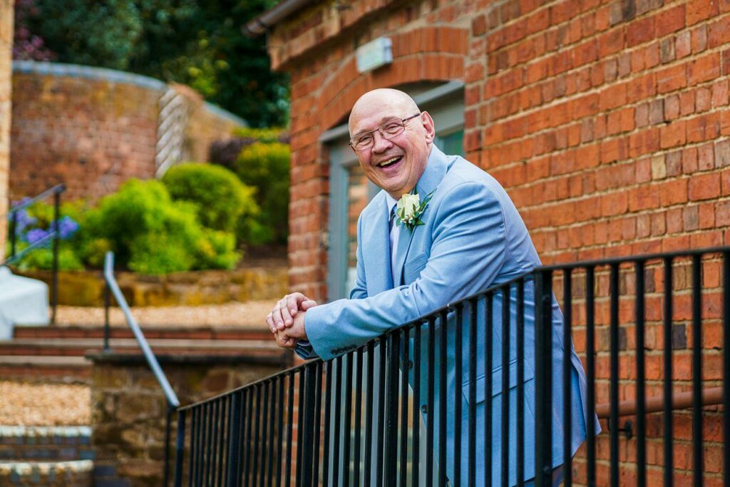 A bald man in a light blue suit leans on a black railing and smiles outside a brick building. Image by Documentary Wedding Photographer Lewis Bishop Photography.