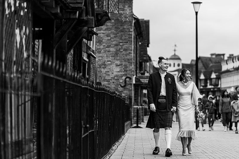 A couple in wedding attire walk hand-in-hand on a street. The man wears a kilt, and the woman wears a white dress. They are smiling and surrounded by buildings and a few other people. Image by Documentary Wedding Photographer Lewis Bishop Photography.